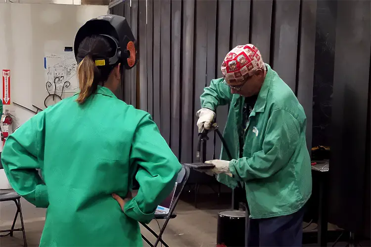 Two people in welding suits and masks, welding at a workbench.